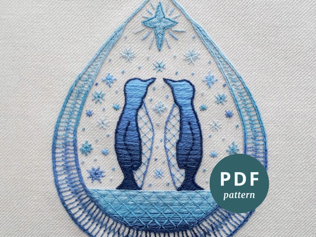 Teardrop shape crewel embroidery design featuring two penguins standing on ice, below a star and surrounded by snowflakes. Stitched in shades of blue and turquoise.