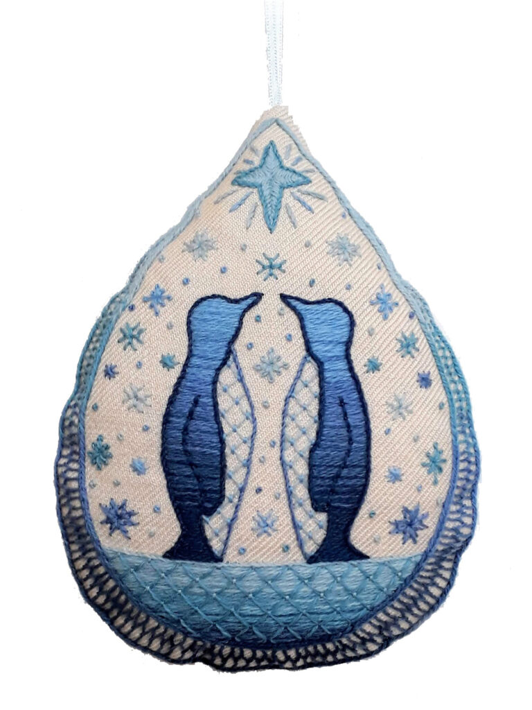 Hanging ornament in a teardrop shape featuring a crewel embroidery design of penguins and snow in shades of blue.