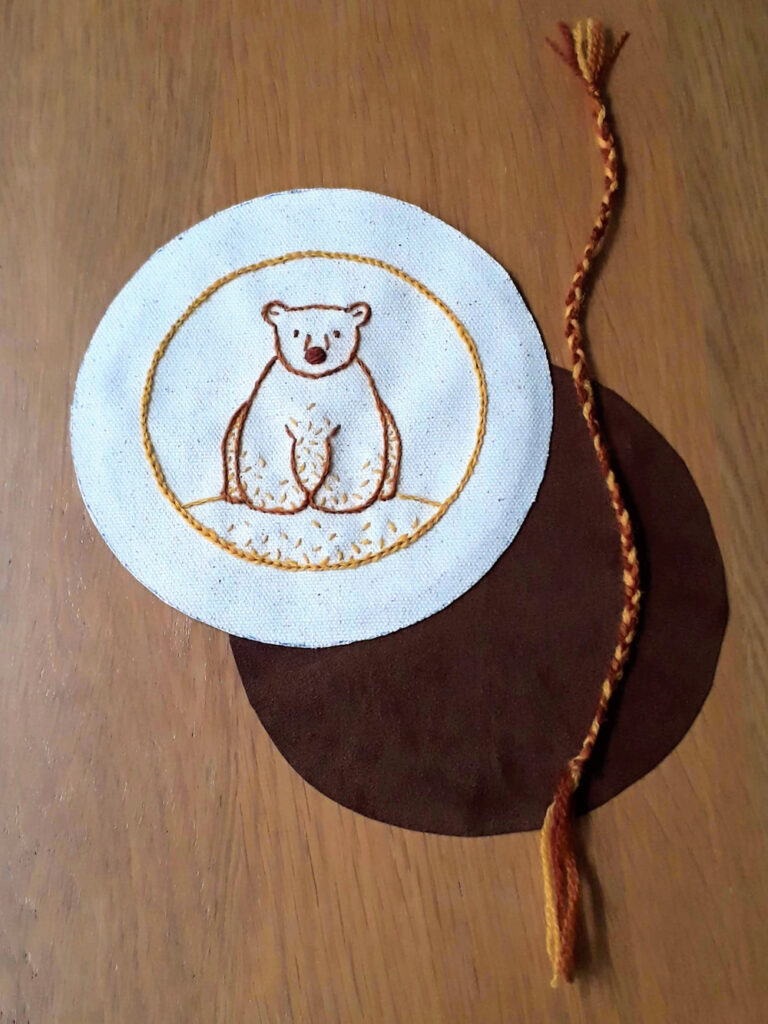 A circle of fabric with an embroidered bear, a circle of brown fabric and a length of braid.