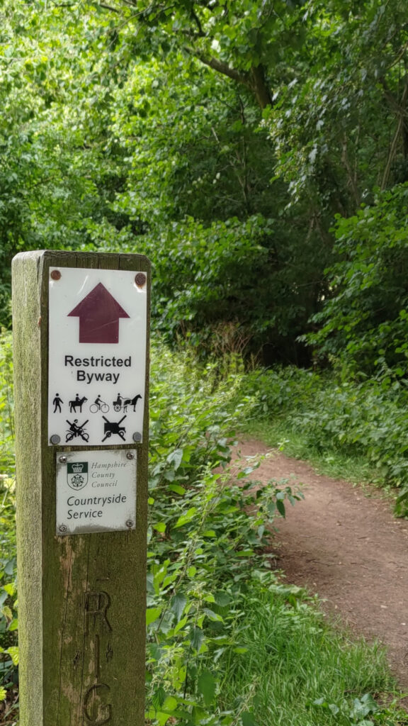 A country walking path surrounding by lots of green. In the foreground on the left is a sign listing people, horses, bicycles and horses and traps as the permitted modes of transport.