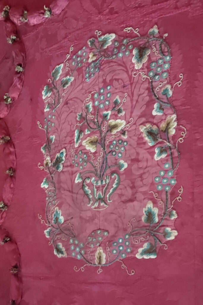 The ceiling of a 4 poster bed canopy featuring rectangular embroidery stitched on pink silk. The embroidery features a central design of stylised flora and fauna, with a border featuring more flora and fauna.