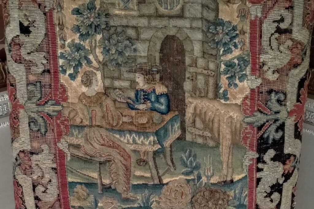 Close up of the embroidery on the back upholstery of an antique chair. The design features a man and a women outside a building.