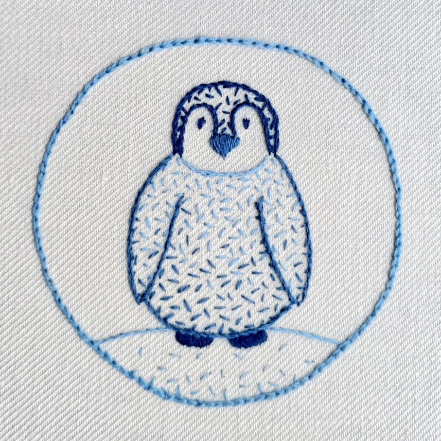 Embroidery of a cute penguin chick standing on ice with a circle border. Stitched in shades of blue.