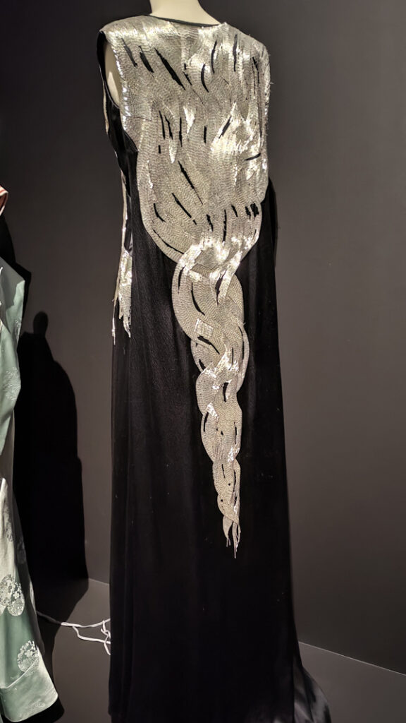 The back of a long, sleeveless black evening dress. Across the shoulders and down the dress is heavy silver embroidery.