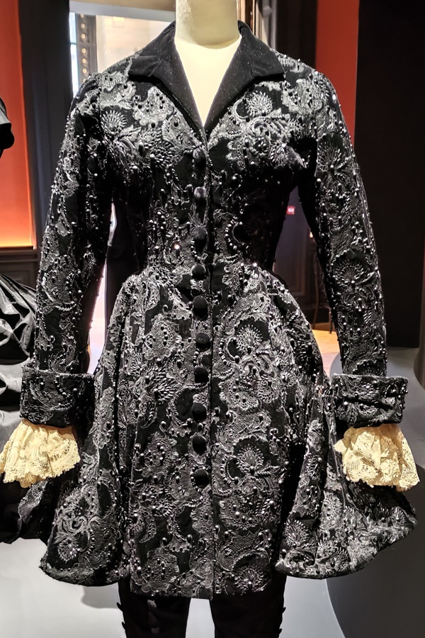 Black couture jacket with embroidery all over it. The bodice is fitted then flares out to knee length. The sleeves are turned up with white ruffles coming out.