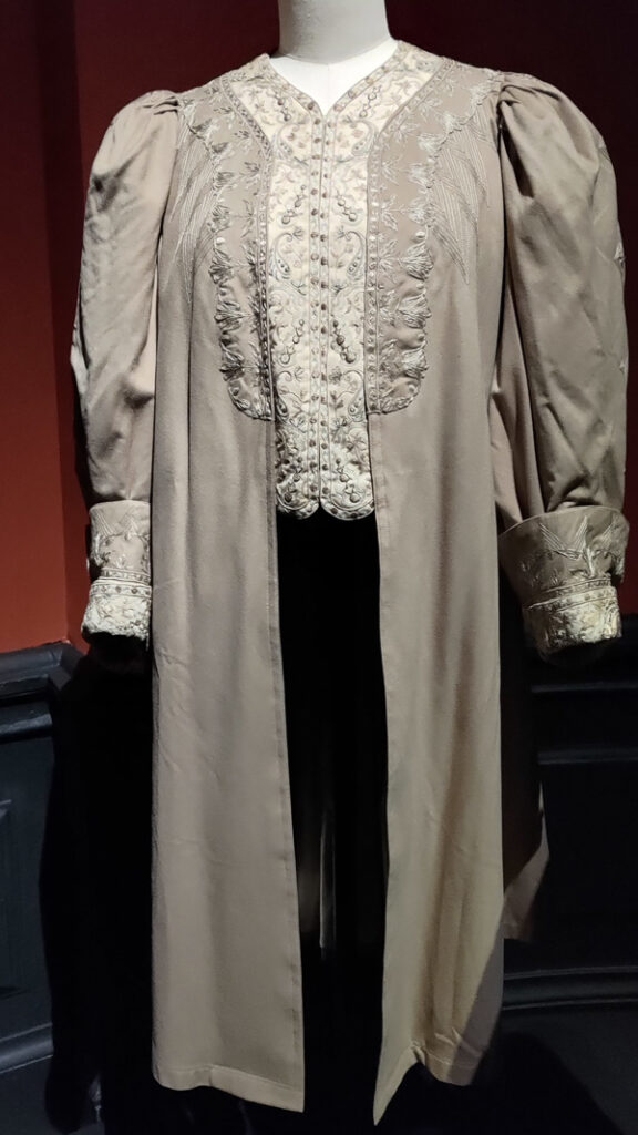 Taupe long coat with lots of fabric gathered into the sleeves. Down the front of the coat is tone on tone embroidery. The cuffs are also embroidered.