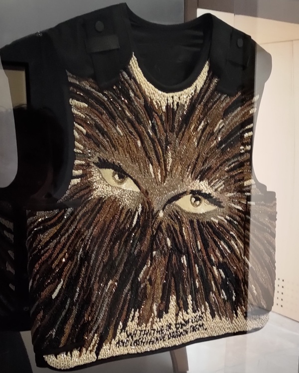 A black military vest that's been heavily embroidered in shades of brown and cream showing a pair of eyes in the centre with fur/hair radiating out.