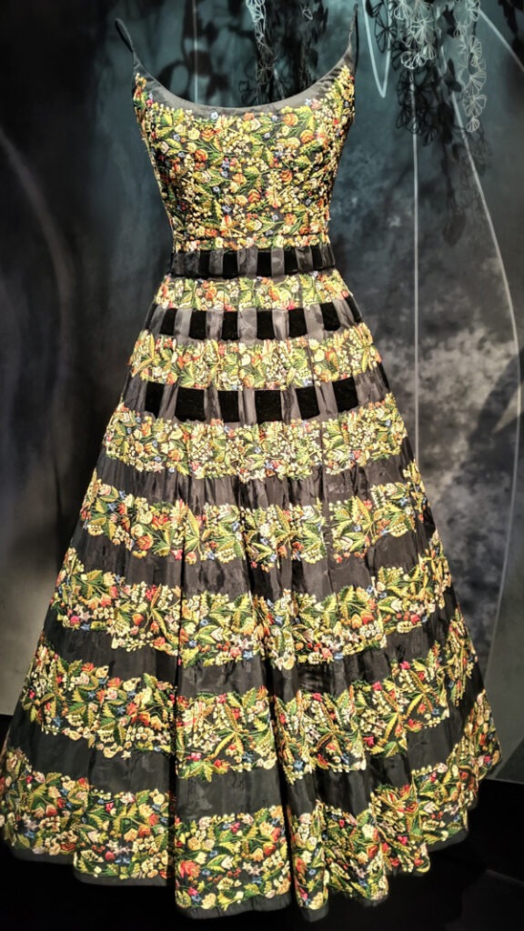 A dress with a fitted bodice and thin straps and a long flared skirt. The bodice is covered with colourful embroidery. The skirt has stripes of colourful embroidery and stripes of the base black fabric.