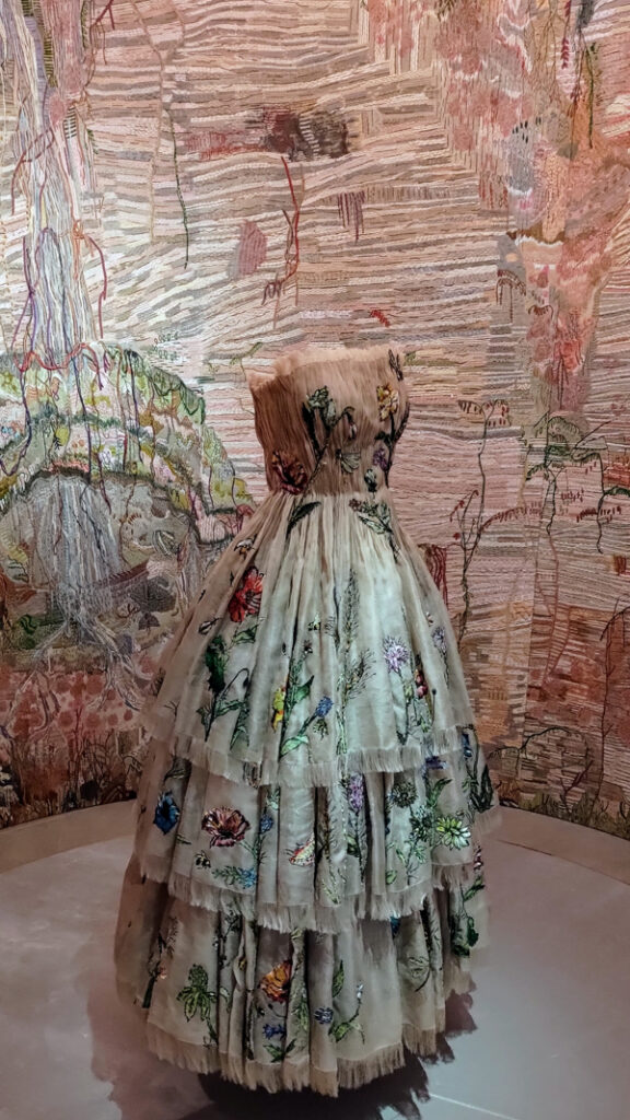 Strapless dress in neutral shades with embroidery all over, some in greens and reds. The background wall is also embroidered to match the dress.