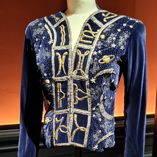 Photo of a hip length evening jacket made of a blue fabric and heavily stitched with gold embroidery showing signs of the zodiac, planets, stars and other astronomical images.