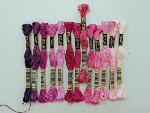 A selection of stranded cotton skeins lined up next to each other, sorted by tone, in shades of purples and pinks.