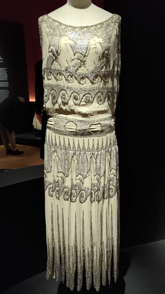 A sleeveless column dress in white with all over silver embroidery. Around the top of the bodice and skirt the embroidery forms the shape of a series of horses.