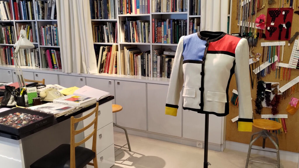 Inside a design studio. The back wall is covered with bookcases and lots of books. In the foreground there is a women's colour blocked jacket in red, blue, yellow, white and black. To the side of the image is the end of a desk.