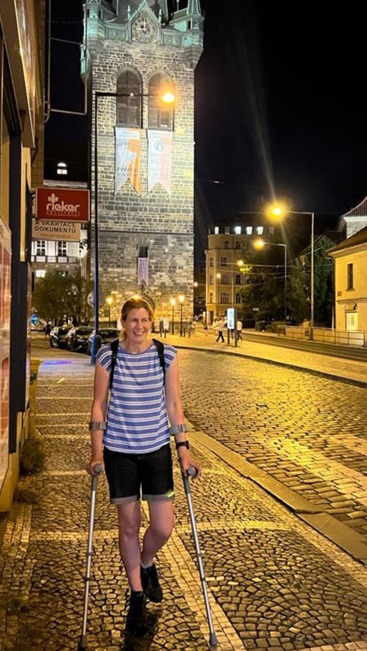 A white woman standing on a cobbled street using crutches at night, with an illuminated tower behind.