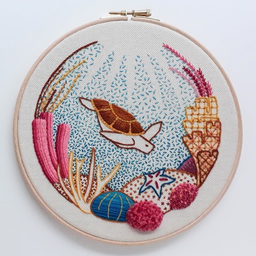 Crewel embroidery of a turtle swimming among coral, mounted in a circle embroidery hoop.