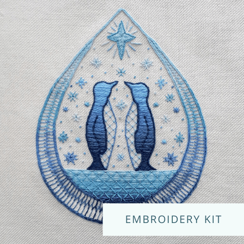 Crewelwork embroidery of two penguins standing on ice surrounded by snowflakes and a star within a wide teardrop shape. Text overlap says "Embroidery kit".
