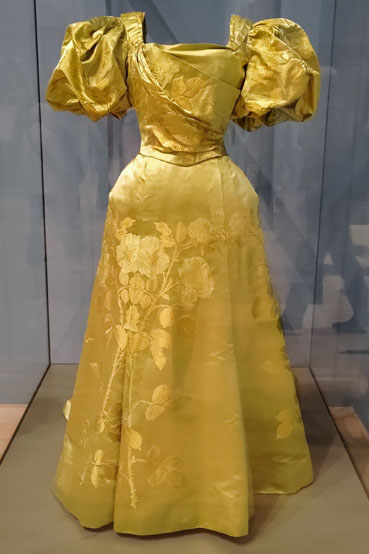 A gold evening dress with draping across the bodice and large elbow length sleeves.