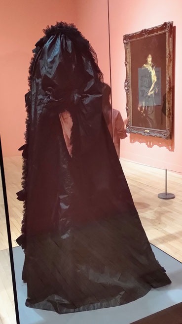 A black opera cloak with pink lining visible at the sleeves in the foreground. In the background is a portrait where the cloak is being worn.