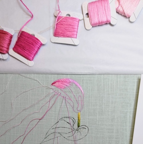 Photo of a partially stitched pink flower. In the top of the image are spools of pink thread ordered by tone. In the bottom of the image is the embroidery with 8 parked threads lined up in order of tone.