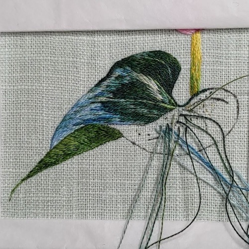 A leaf half stitched in silk shading embroidery with various shades of greens, blues and greys, so that the leaf looks like it's curving.