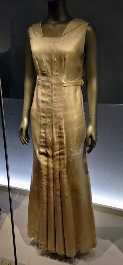 Photo of a light gold dress with a square and folded neckline, and pleats and seams down the front that look like a classical architecture column.