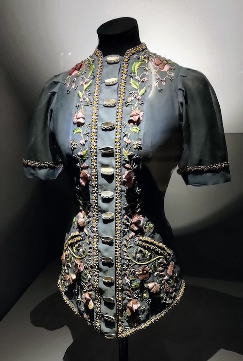 Photo of short sleeve jacket in a light fabric. The front is heavily embellished with floral embroidery and tambourine buttons.