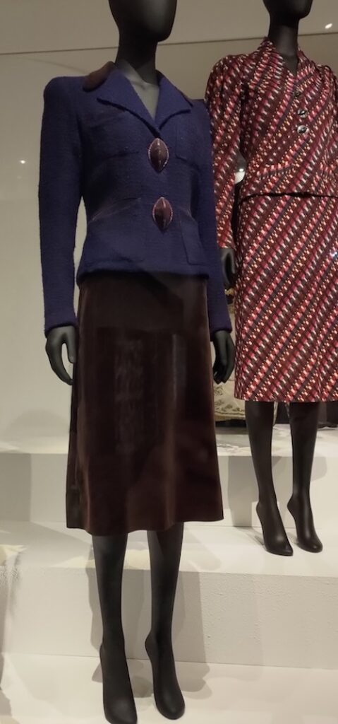 Photo of a skirt suit with A-line brown skirt and blue jacket featuring two very large buttons. In the background is another suit in shades of burgundy.